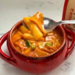 Chewy rice and fish cakes bathed in a fiery gochujang sauce, garnished with and green onions and served on a red bowl.