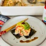 A delectable serving of crab and salmon sushi bake nestled on a bed of seaweed sheets, adorned with creamy avocado slices and a drizzle of spicy sriracha and eel sauce, offering a fusion of flavors and textures.