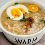 Rotisserie Chicken Arroz Caldo topped with open faced egg and a slice of calamansi and garlic tidbits on a white bowl.