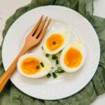 Open-faced 7-minute egg on a white plate with spoon on top of a white table with dark green napkin underneath.