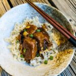 Aromatic beef pares served on a rustic noodle bowl, showcasing tender beef chucks simmered in savory sauce on top squiggly noodles with chopped green onions as a garnish.