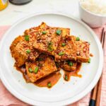 Golden brown crunchy tofu slices drizzled with savory soy glaze, arranged on a white plate, offering a delightful combination of textures and flavors.