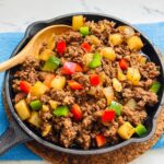 Ground beef stir fry with small cubed potatoes and bell peppers on a black iron pan with wooden spoon inside.