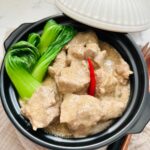 Spicy pork with creamy coconut sauce with bok choy and thai chili in a black claypot with lid on the side.