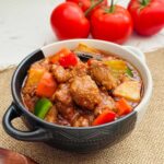Pork menudo in a black and white bowl with some tomatoes on the side.