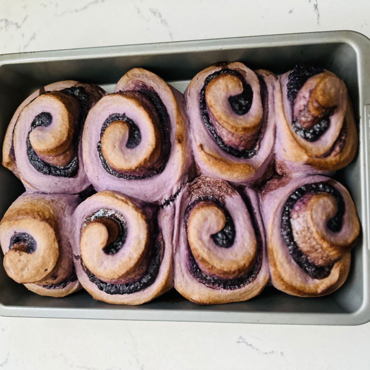 Close-up of freshly baked Ube Rolls on a cooling rack. The rolls have a golden-brown exterior and a soft, fluffy interior with a vibrant purple color, showcasing their delicious texture and aroma right out of the oven.