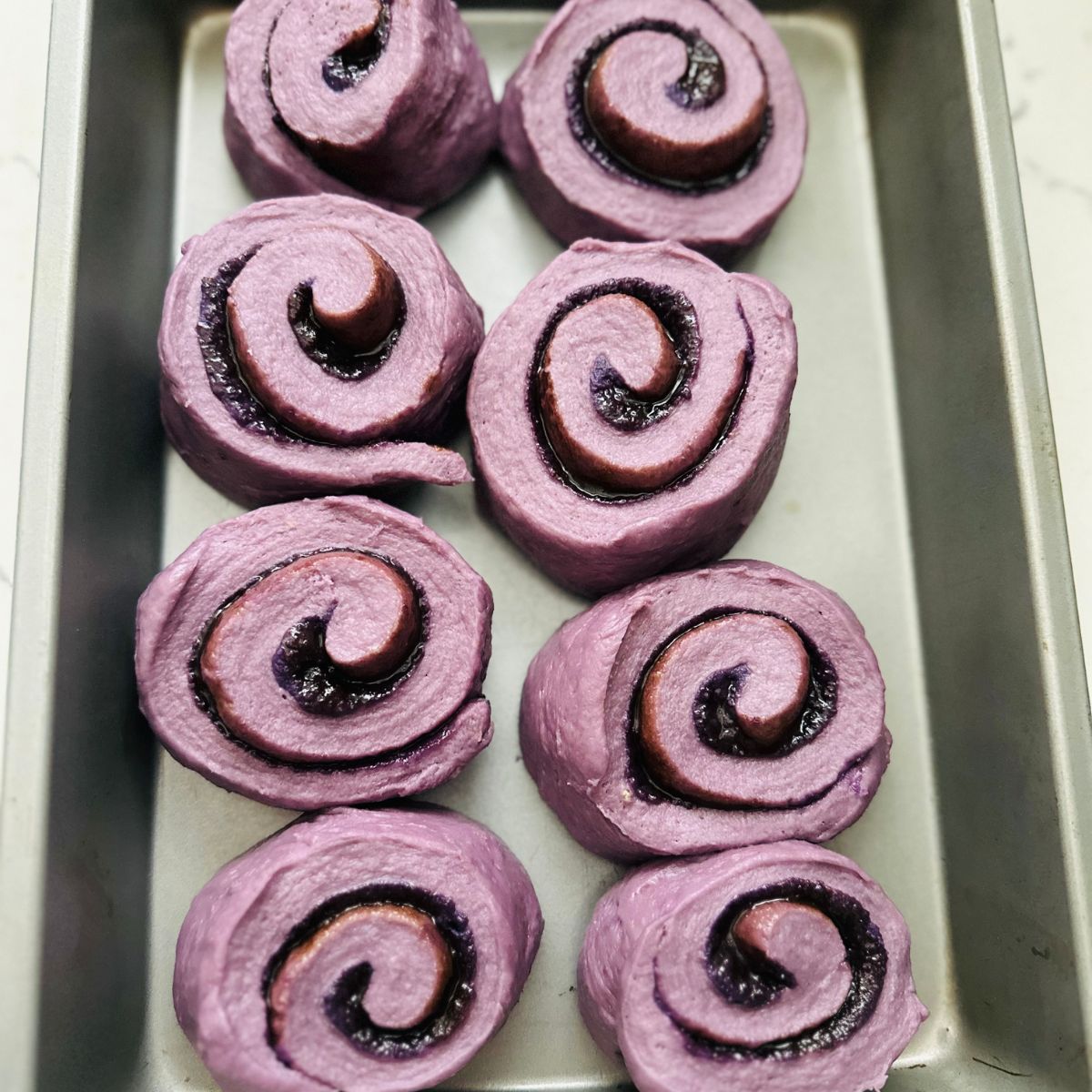 Ube Rolls on a baking tray, ready for their second proofing. The rolls are arranged on the tray, showing their soft, puffy texture and vibrant purple color, prepared for their final rise before baking.