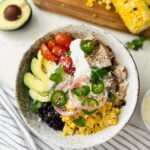 A rice bowl being layered with cooked rice, black beans, corn, avocado, tomatoes, and onions topped with sliced chicken, cotija cheese, sour cream, and cilantro.