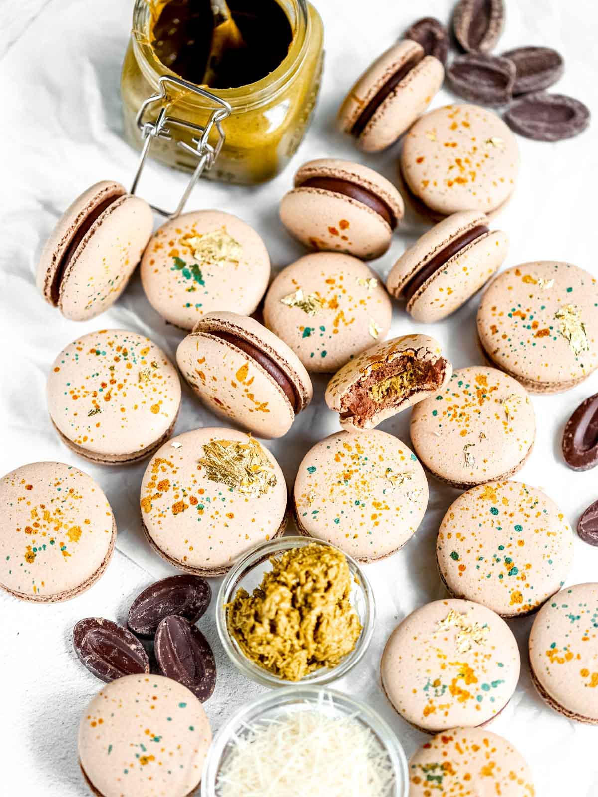 Delicate Dubai chocolate macarons filled with pistachio ganache and dusted with edible gold, styled on a marble surface.