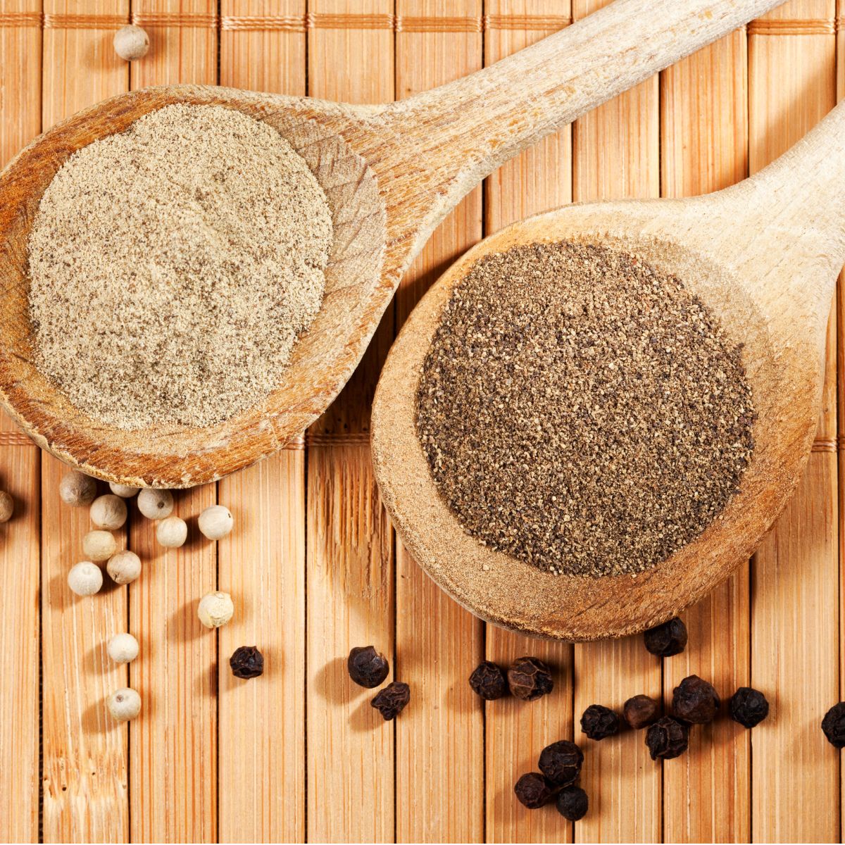 Ground white pepper and black pepper in wooden spoons with whole peppercorns on a bamboo surface.