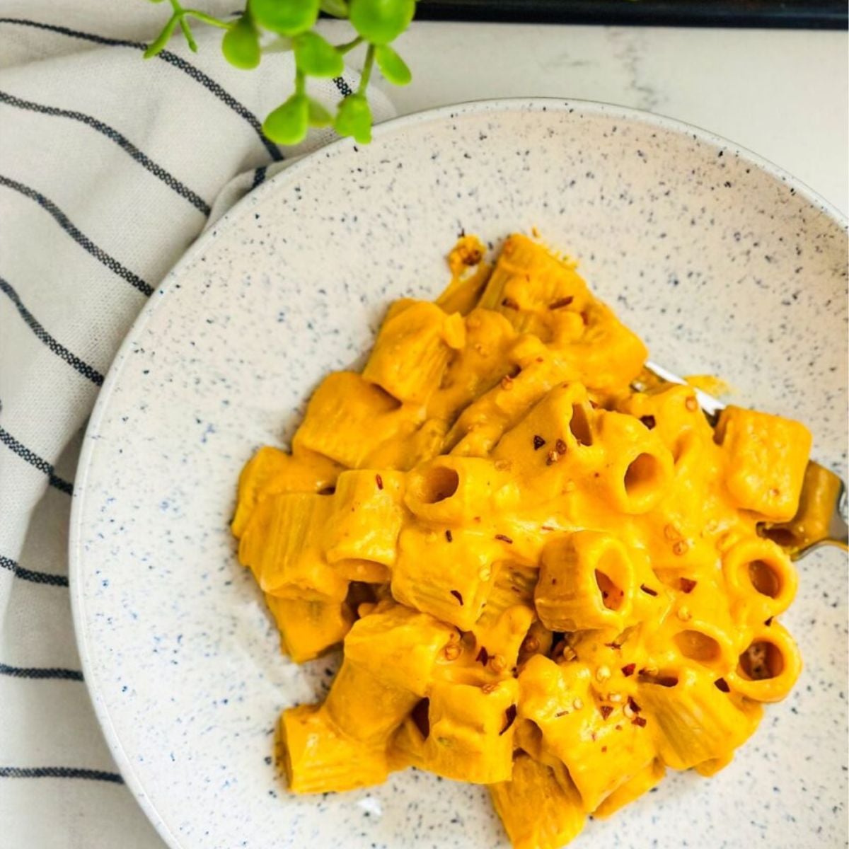 Creamy pumpkin pasta with cream cheese sauce and red pepper flakes served on a speckled plate.