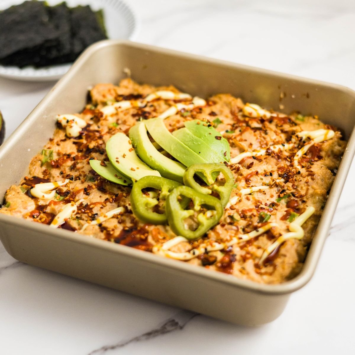 Pan of spicy tuna sushi bake made with rice, furikake, and baked tuna mixture, ready to serve and topped with japapeno and sliced avocados.