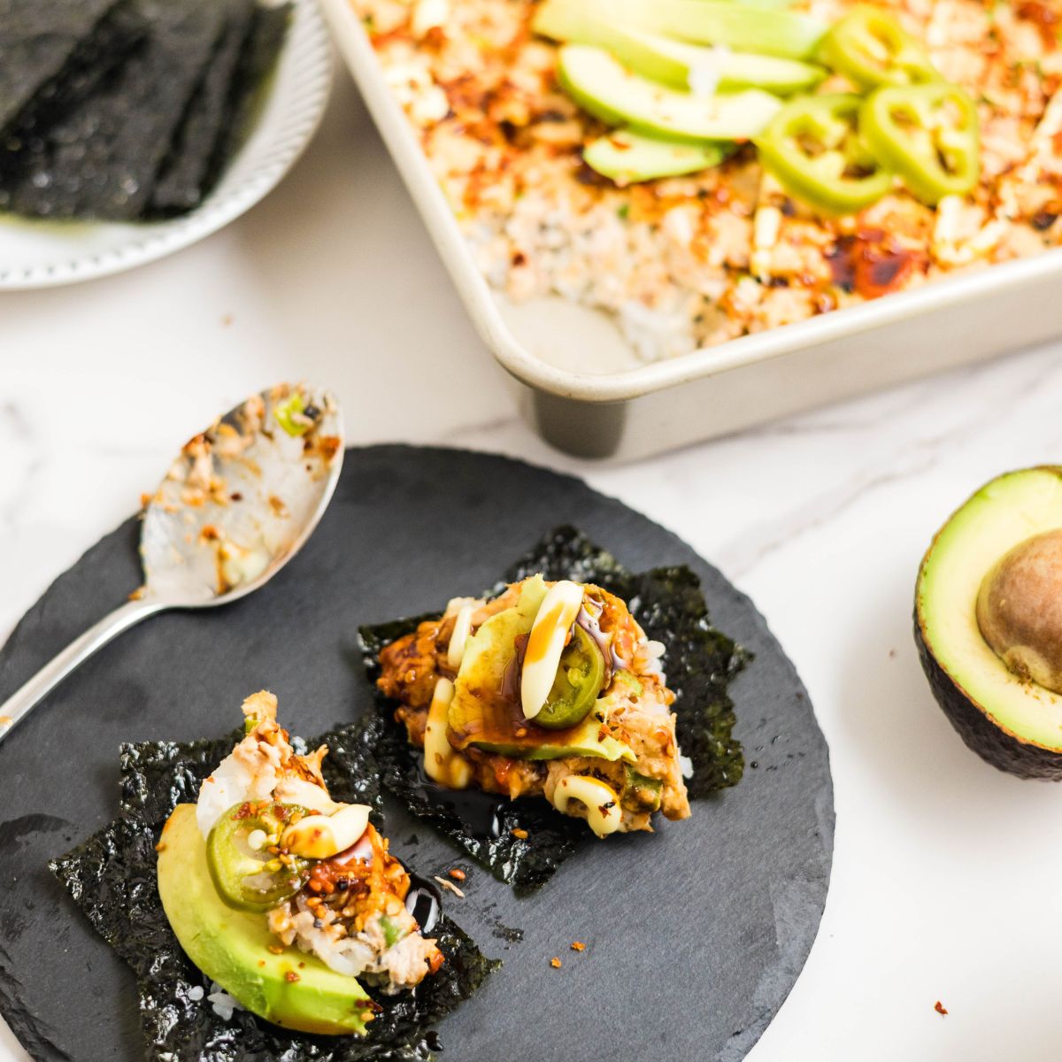 Spoonful of spicy tuna sushi bake served on seaweed with avocado and drizzled with eel sauce, sriracha, and mayo.