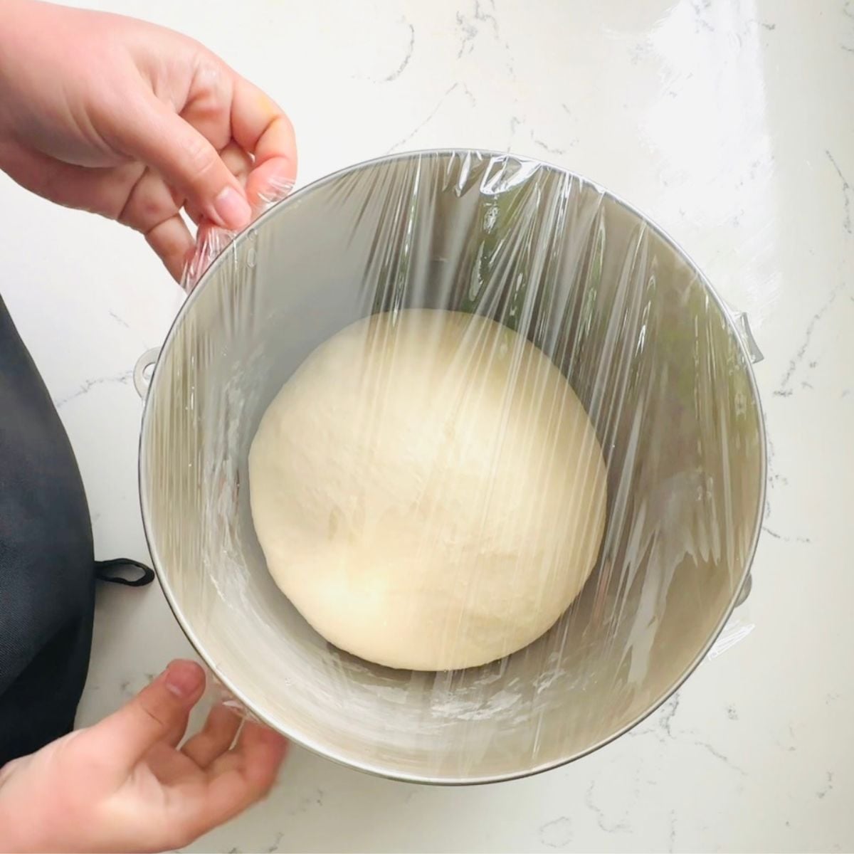 Smooth ball of dough resting in a metal mixing bowl covered tightly with plastic wrap for proofing.