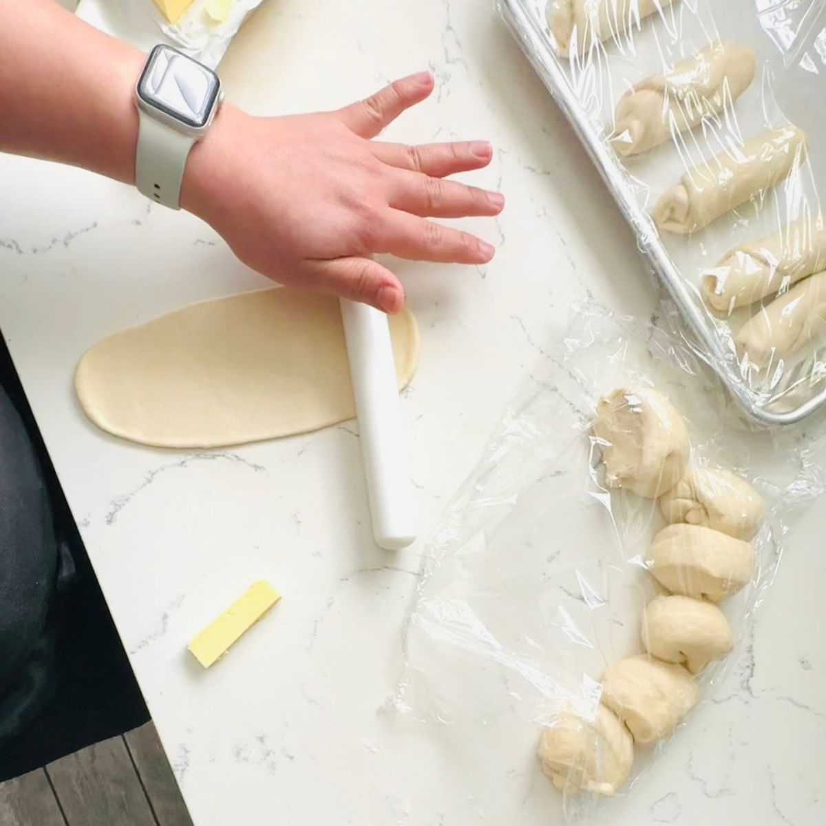 Hand rolling out shio pan dough into an oval on a white countertop, with butter pieces and shaped dough portions nearby.