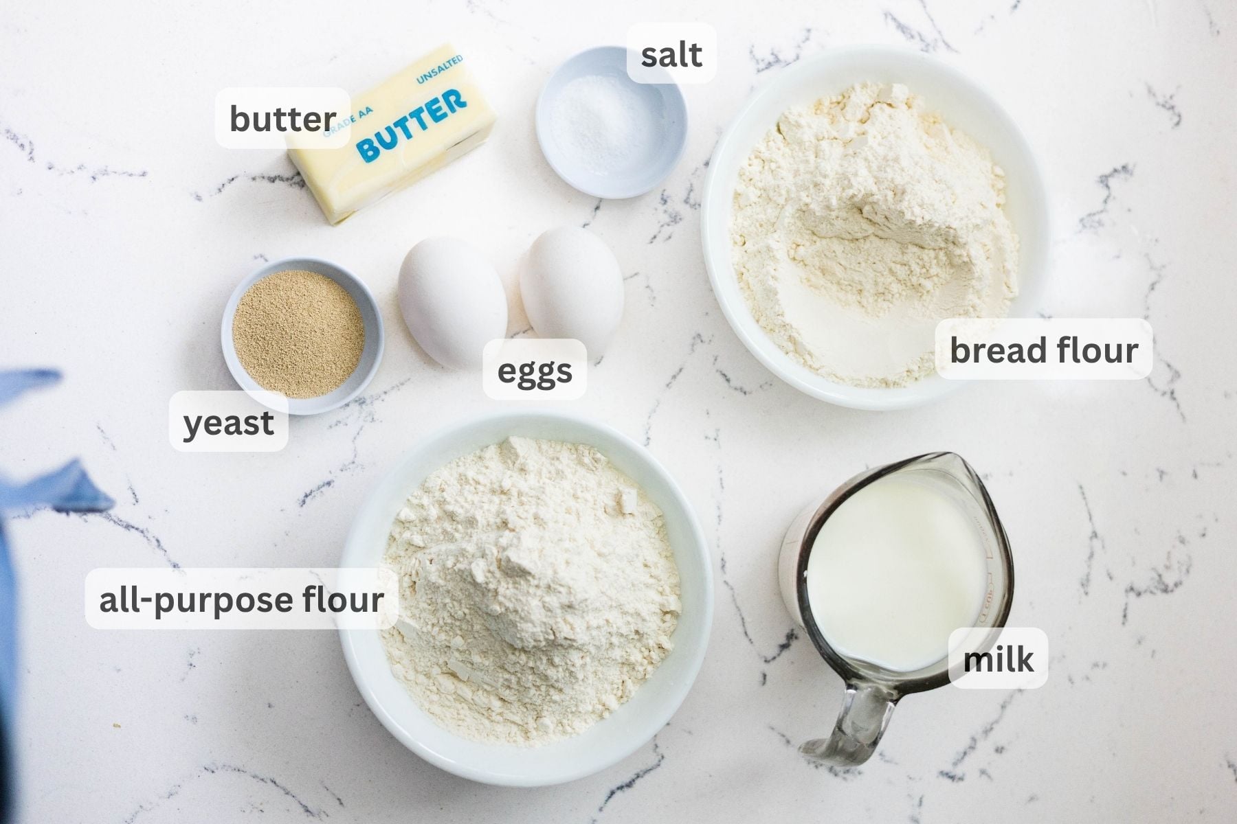 Overhead view of cinnamon roll ingredients on a white countertop including butter, salt, bread flour, yeast, eggs, all purpose flour, and milk.