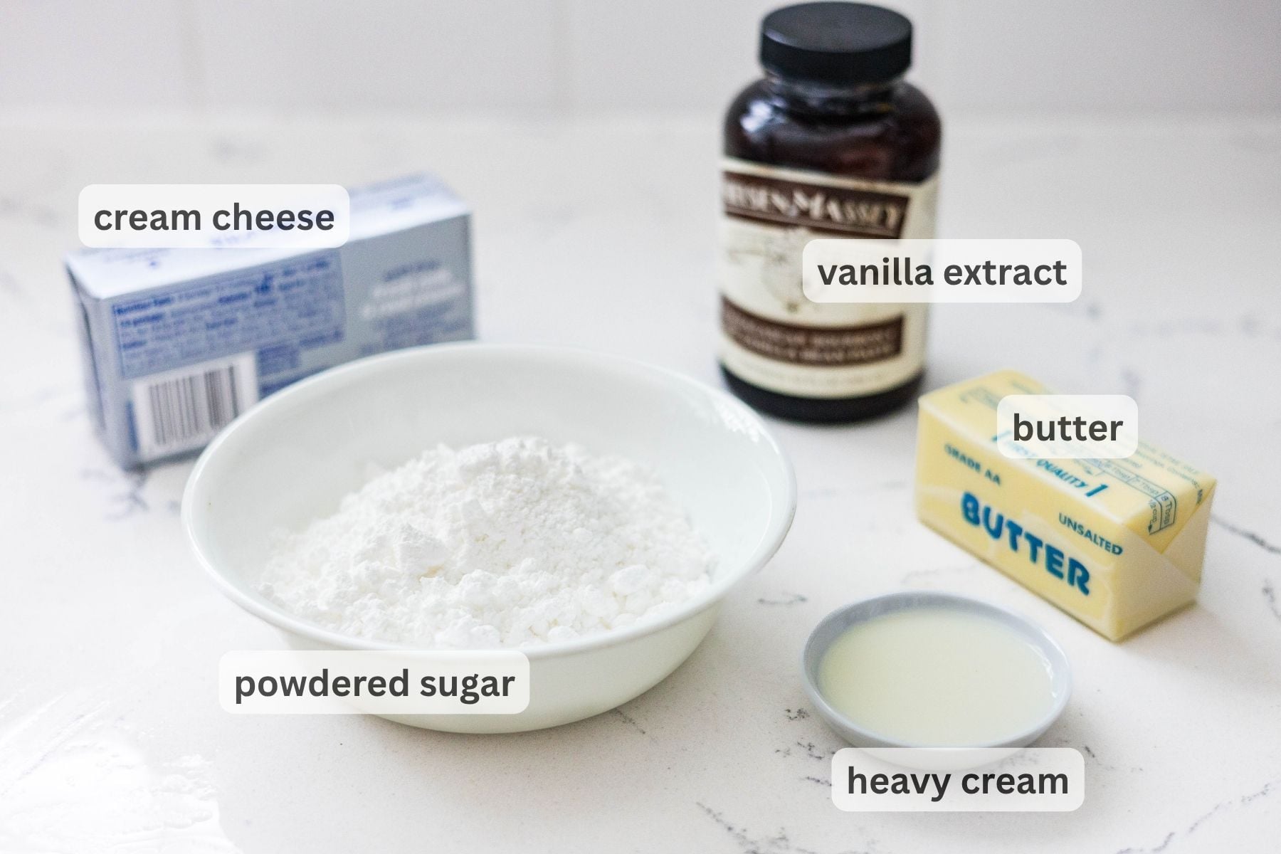 Overhead view of cream cheese frosting ingredients on a white countertop including cream cheese, vanilla extract, butter, powdered sugar, and heavy cream.