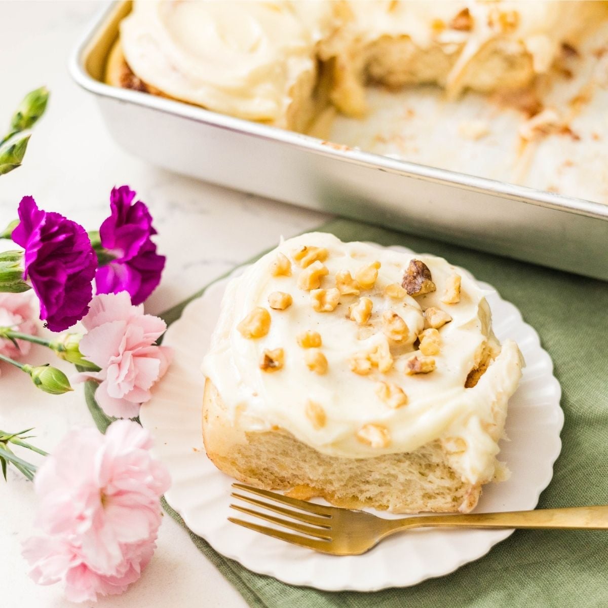 Classic cinnamon roll with cream cheese frosting on a white plate topped with chopped nuts beside a baking pan