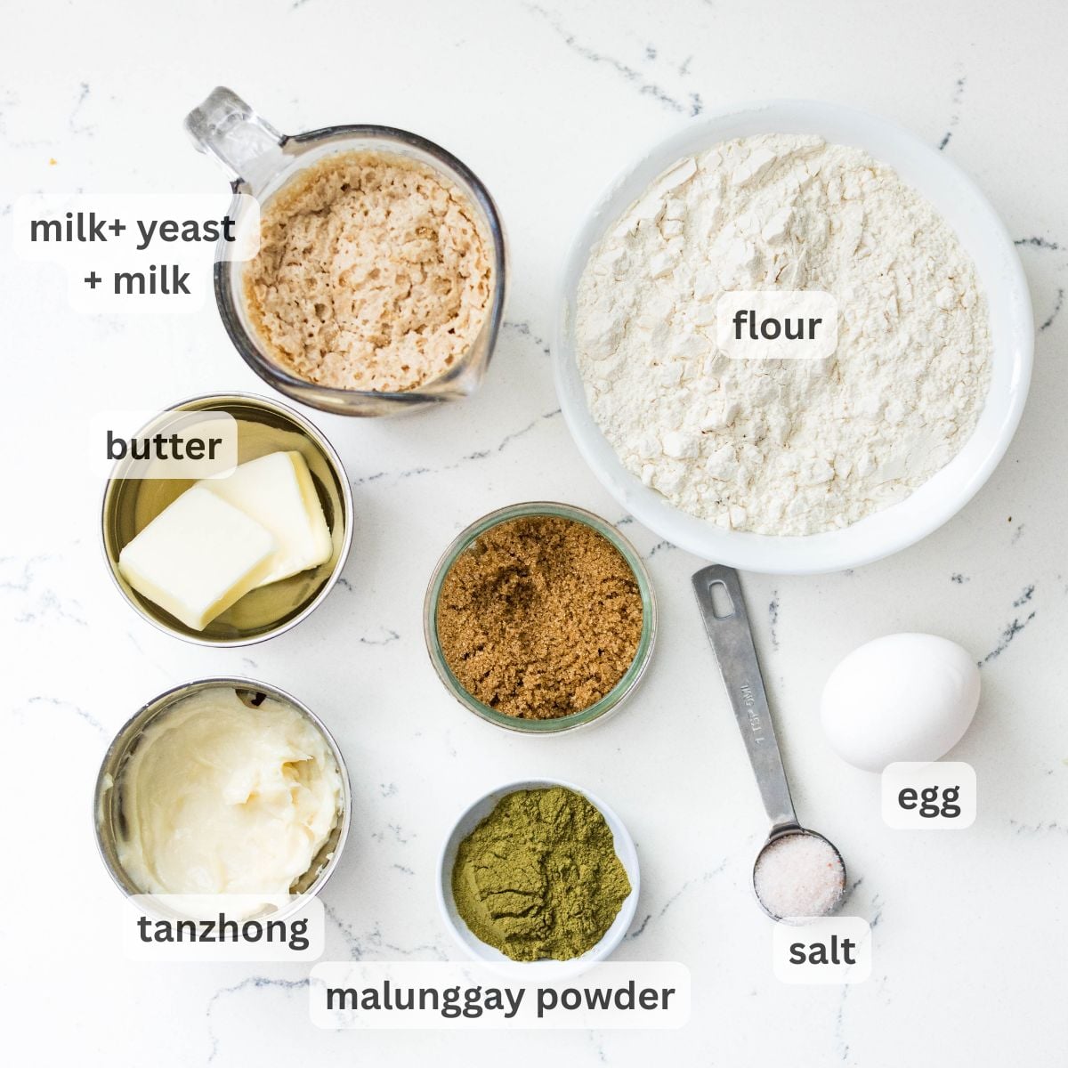 Ingredients for malunggay pandesal including flour, breadcrumbs, butter, mashed potato, brown sugar, egg, salt, and malunggay powder on a white counter.