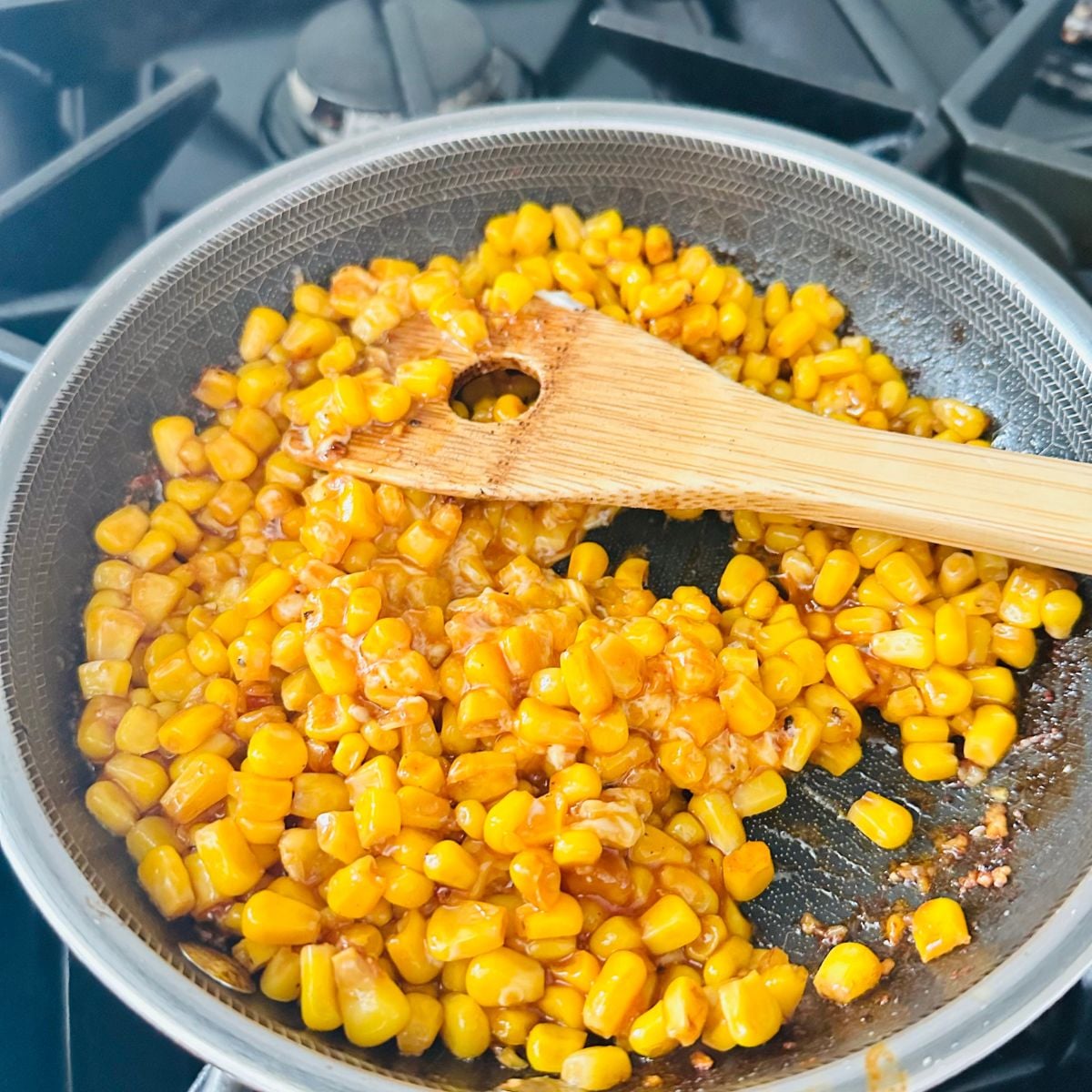 Sweet corn cooking in a skillet with sauce and a wooden spatula.