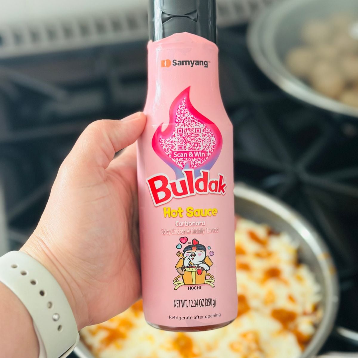 Hand holding a bottle of Buldak Carbonara hot sauce over a pan of cheesy corn.