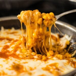 Spoon lifting cheesy Korean corn with long melted cheese pulls from the skillet.