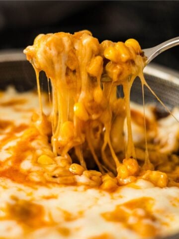 Spoon lifting cheesy Korean corn with long melted cheese pulls from the skillet.