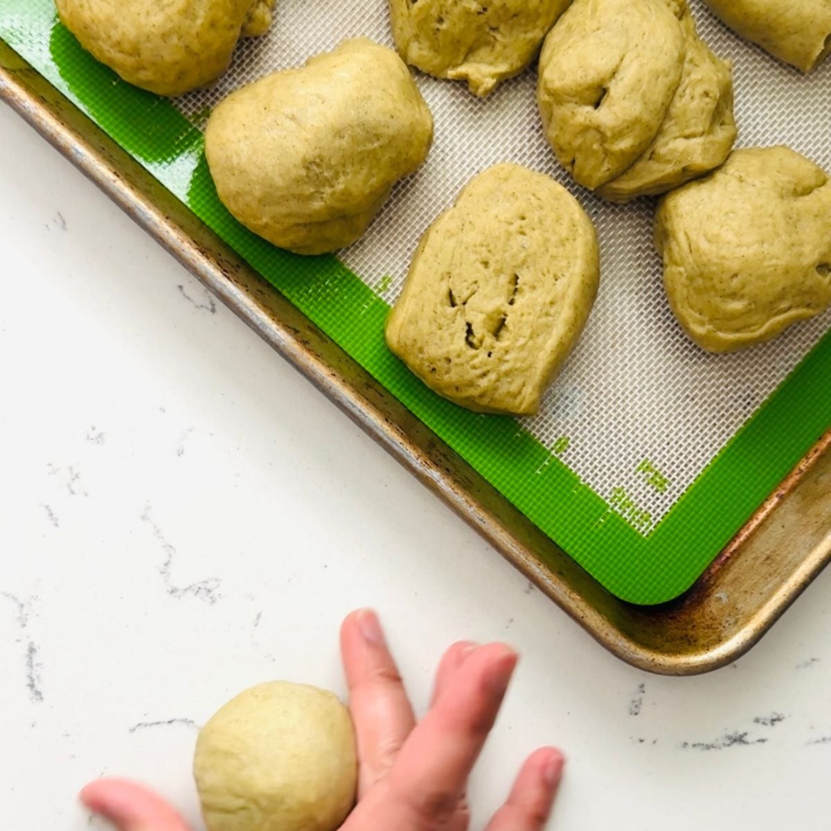 Shaped malunggay pandesal dough portions on a baking sheet with one dough ball being rolled by hand.