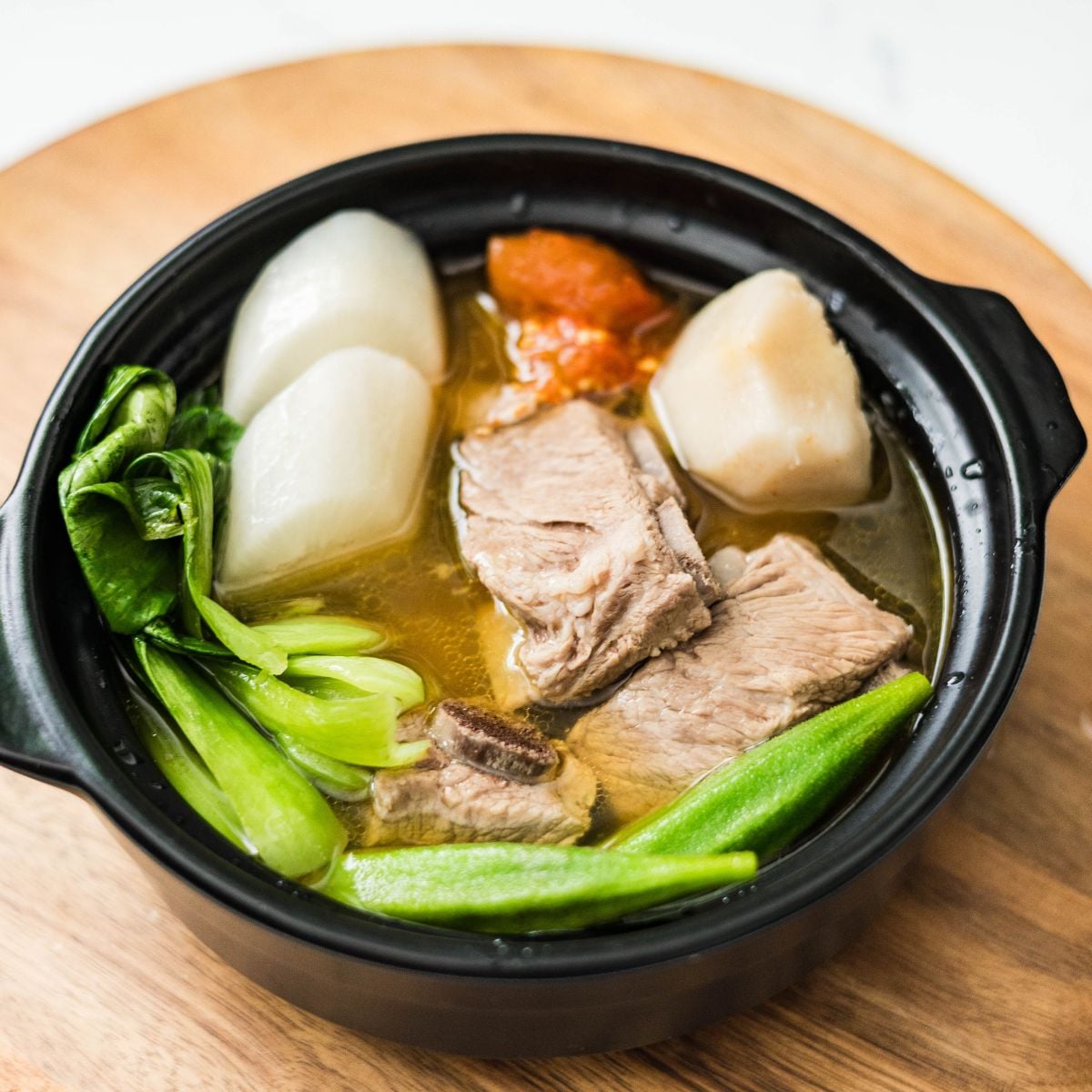 Pork sinigang with spare ribs served in a black bowl with radish, taro, bok choy, okra, and tomato.