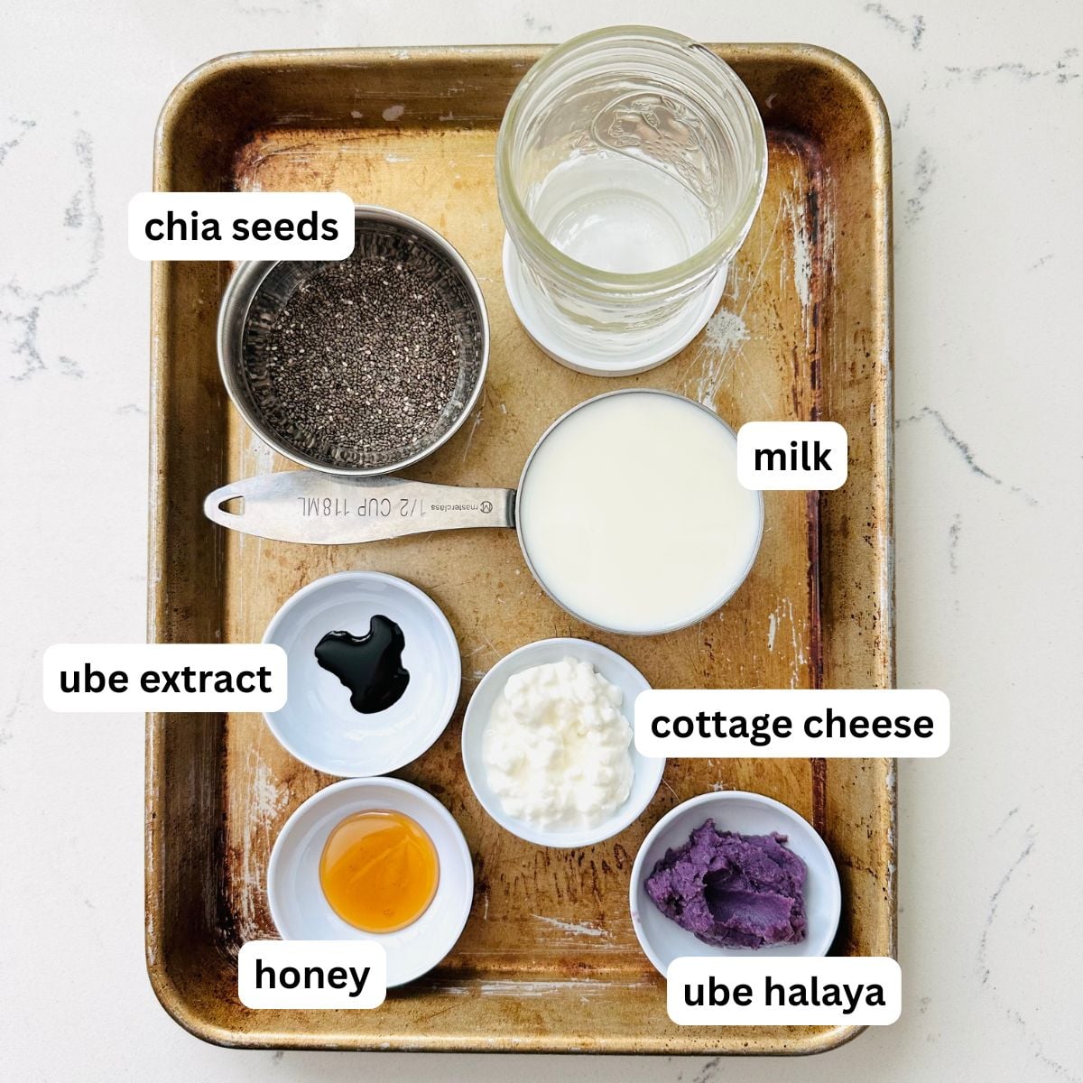 Ingredients for ube chia pudding on a baking sheet including chia seeds, milk, cottage cheese, ube halaya, honey, and ube extract