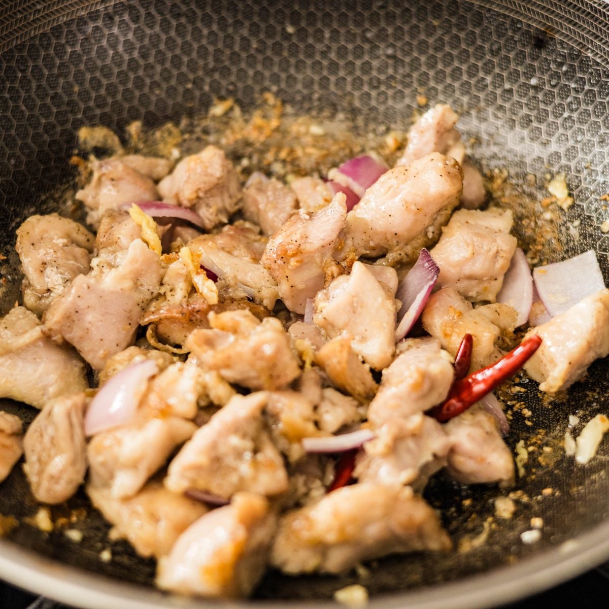 Bite-sized chicken pieces cooking in a wok with garlic, red onion, and dried chilies.