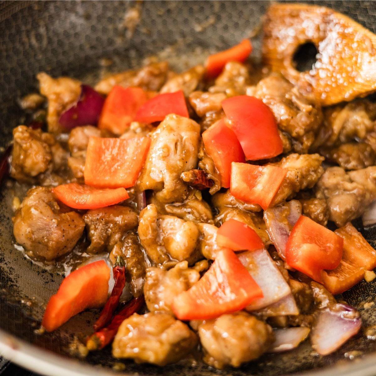 Bite-sized chicken pieces cooking in a wok with garlic, red onion, and dried chilies.