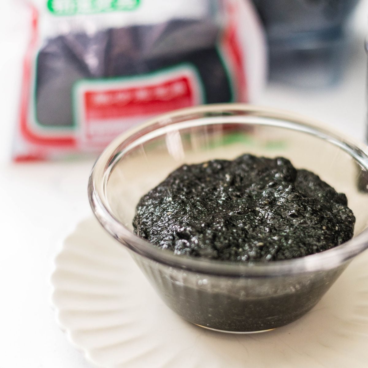 Homemade black sesame paste in a glass bowl with black sesame seeds in the background.