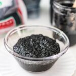 Homemade black sesame paste in a glass bowl with black sesame seeds in the background.