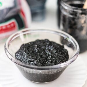 Homemade black sesame paste in a glass bowl with black sesame seeds in the background.