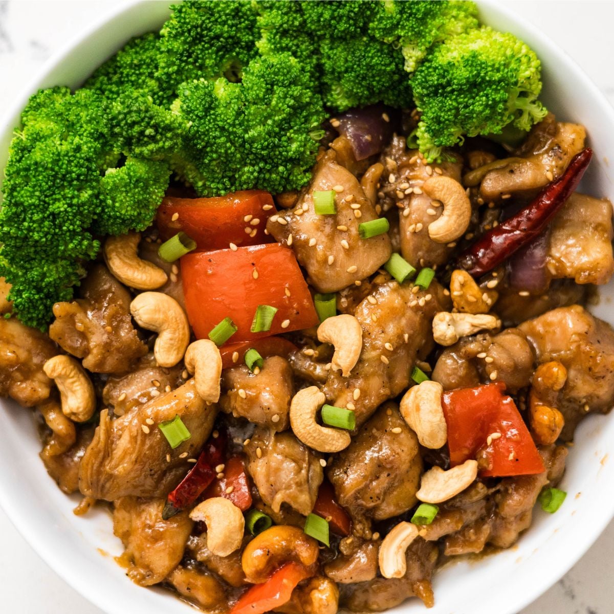 Cashew chicken bowl with steamed broccoli, red bell peppers, dried chilies, sesame seeds, and chopped green onions.