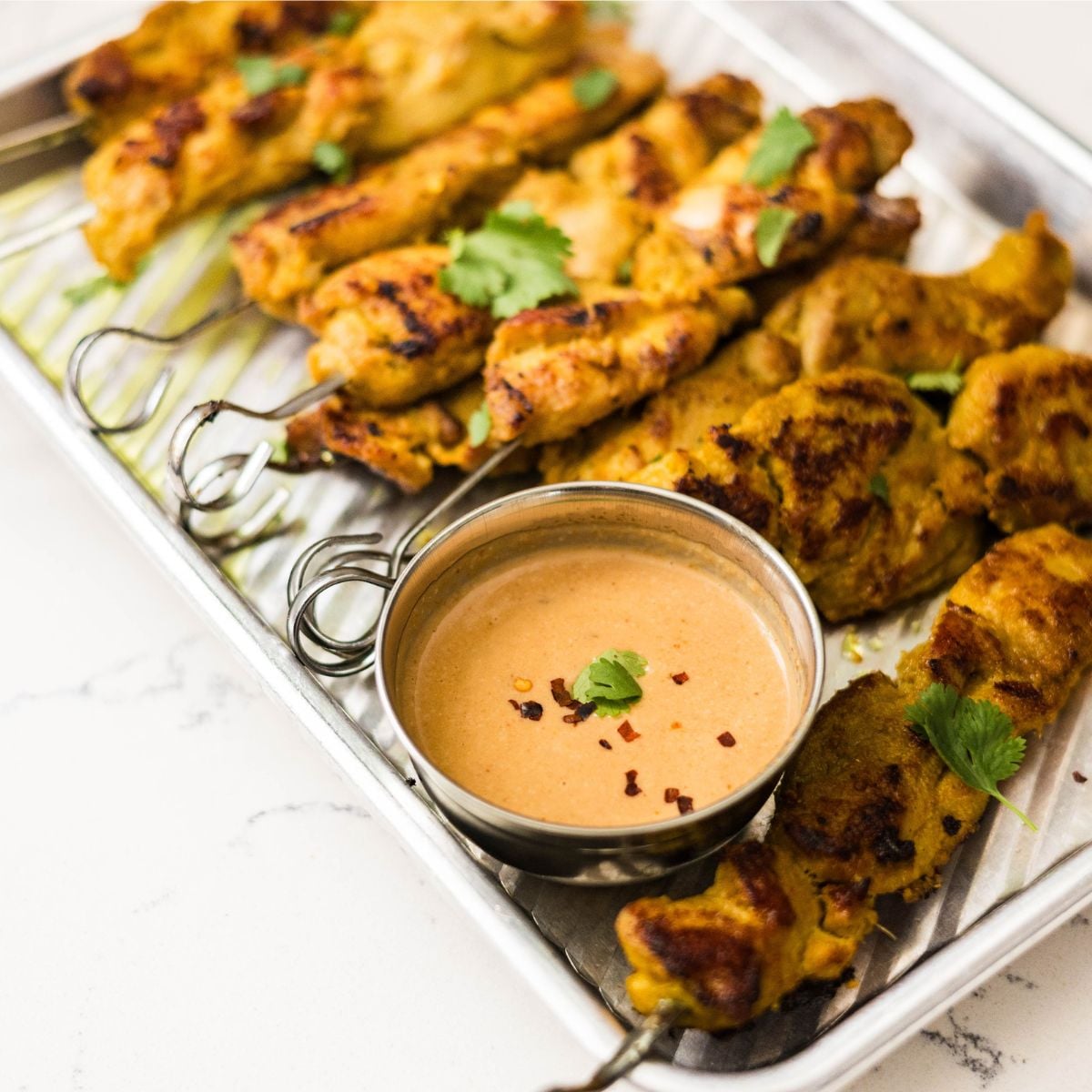 Thai chicken satay skewers served on a metal tray with sliced cucumbers, cilantro, peanut sauce, and nam jim dipping sauce.