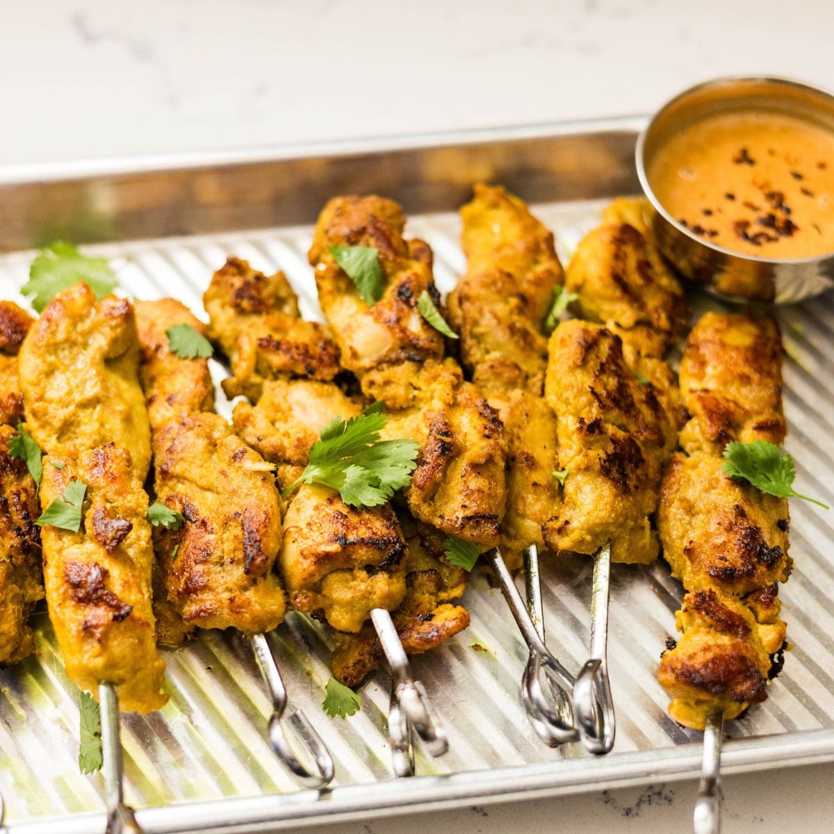 Thai chicken satay skewers served on a metal tray with sliced cucumbers, cilantro, and peanut sauce.