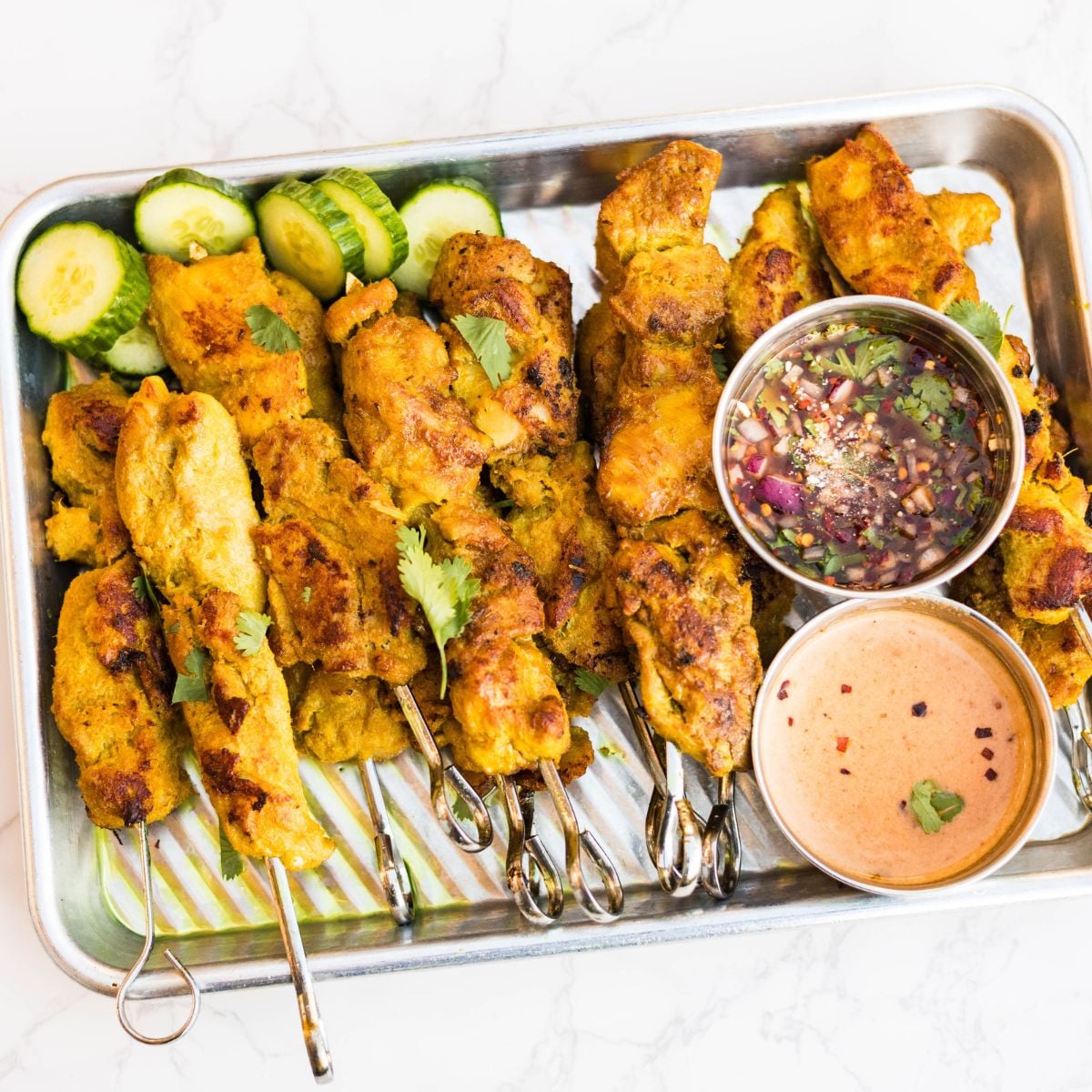 Thai chicken satay skewers served on a metal tray with sliced cucumbers, cilantro, peanut sauce, and nam jim dipping sauce.