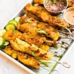 Thai chicken satay skewers served on a metal tray with sliced cucumbers, cilantro and nam jim dipping sauce.