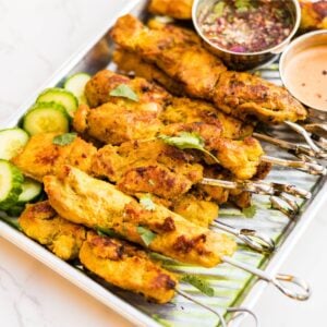 Thai chicken satay skewers served on a metal tray with sliced cucumbers, cilantro and nam jim dipping sauce.