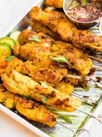 Thai chicken satay skewers served on a metal tray with sliced cucumbers, cilantro and nam jim dipping sauce.