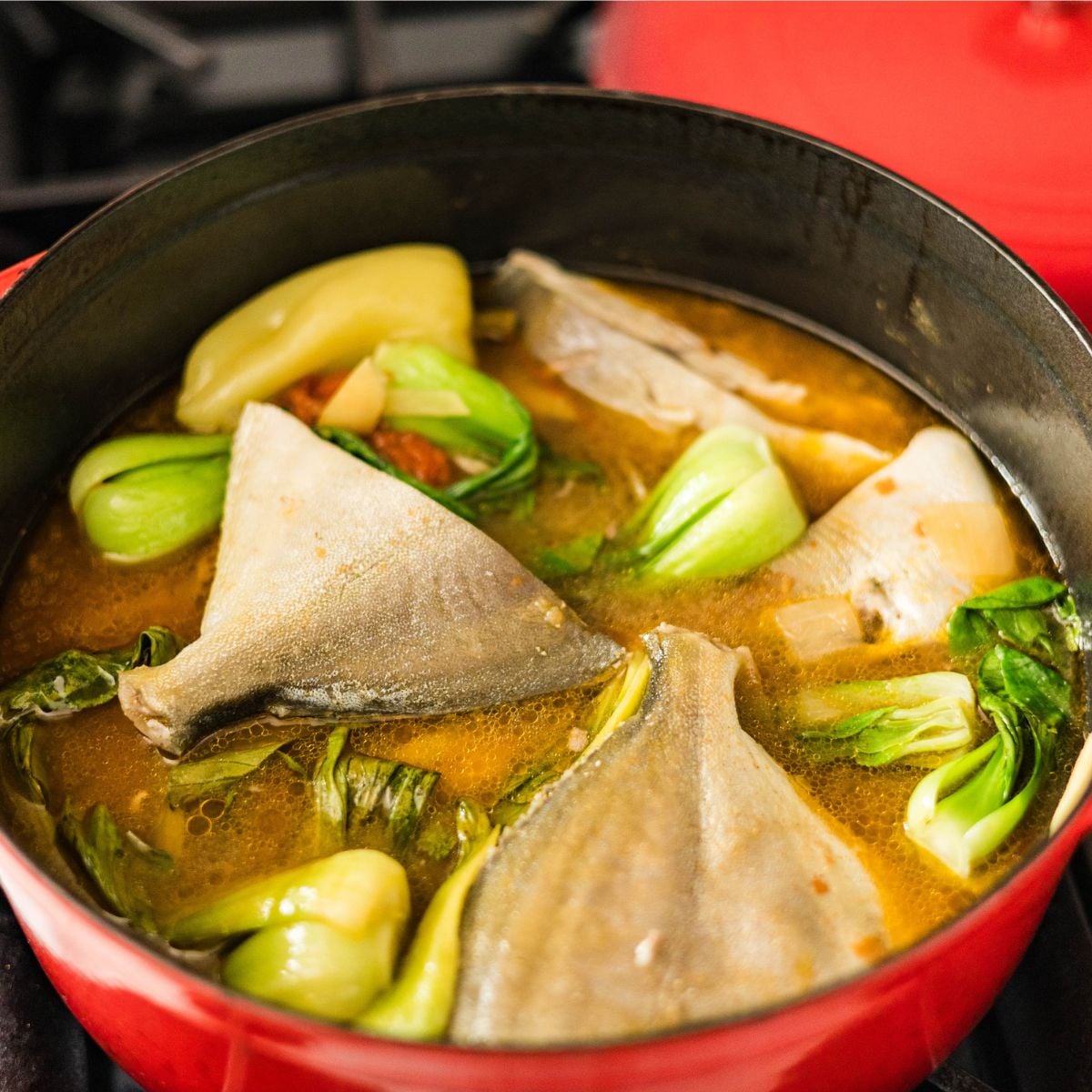 Pompano sinigang sa miso simmering in a red pot with bok choy, tomatoes, ginger, and a savory tamarind miso broth.