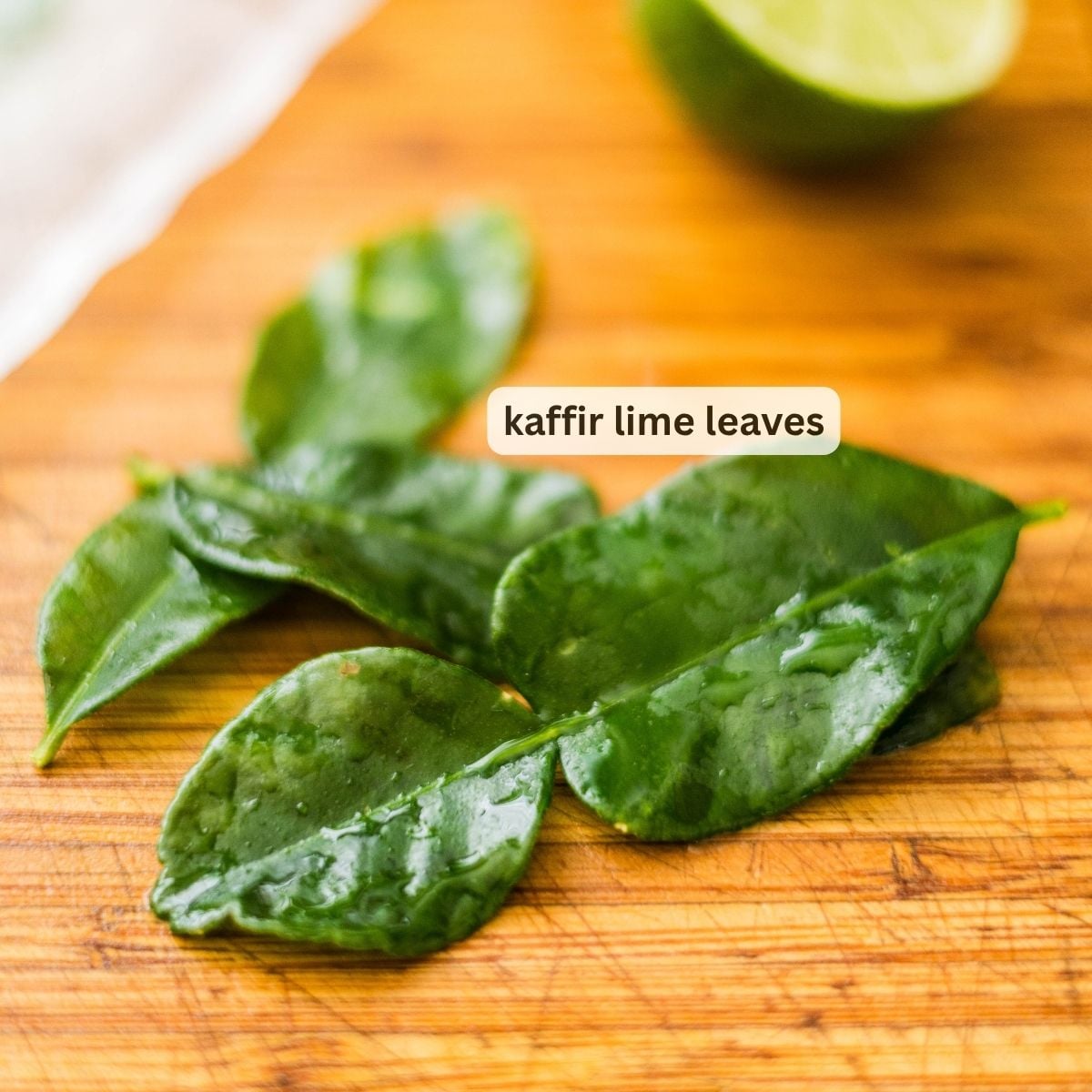 Kaffir lime leaves on a wooden cutting board with a sliced lime in the background.