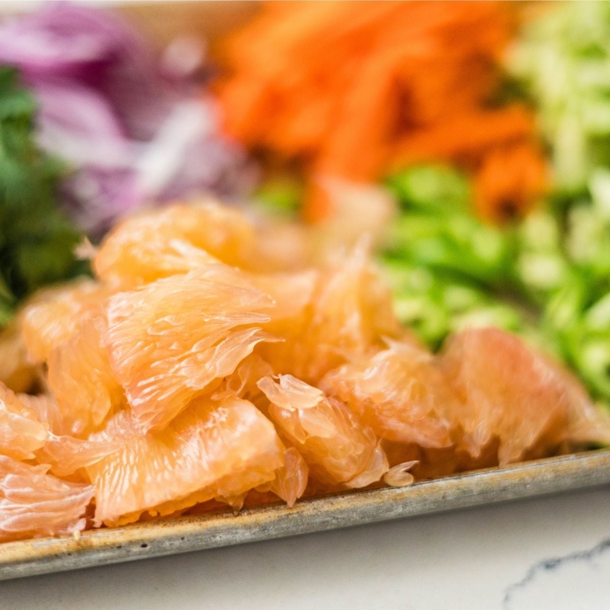 Pomelo segments on a tray with chopped vegetables blurred in the background.