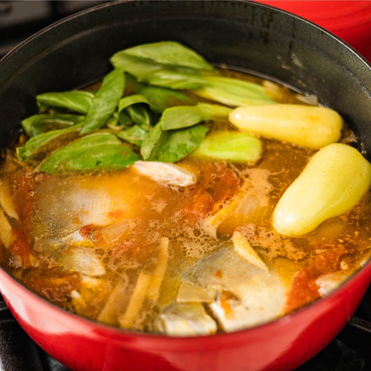 Pompano sinigang sa miso simmering in a red pot with bok choy, tomatoes, ginger, and a savory tamarind miso broth.