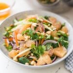 Pomelo salad in a bowl with fresh herbs, cucumber, carrots, red onion, chili, peanuts, and a wooden spoon nearby.