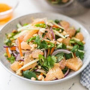 Pomelo salad in a bowl with fresh herbs, cucumber, carrots, red onion, chili, peanuts, and a wooden spoon nearby.