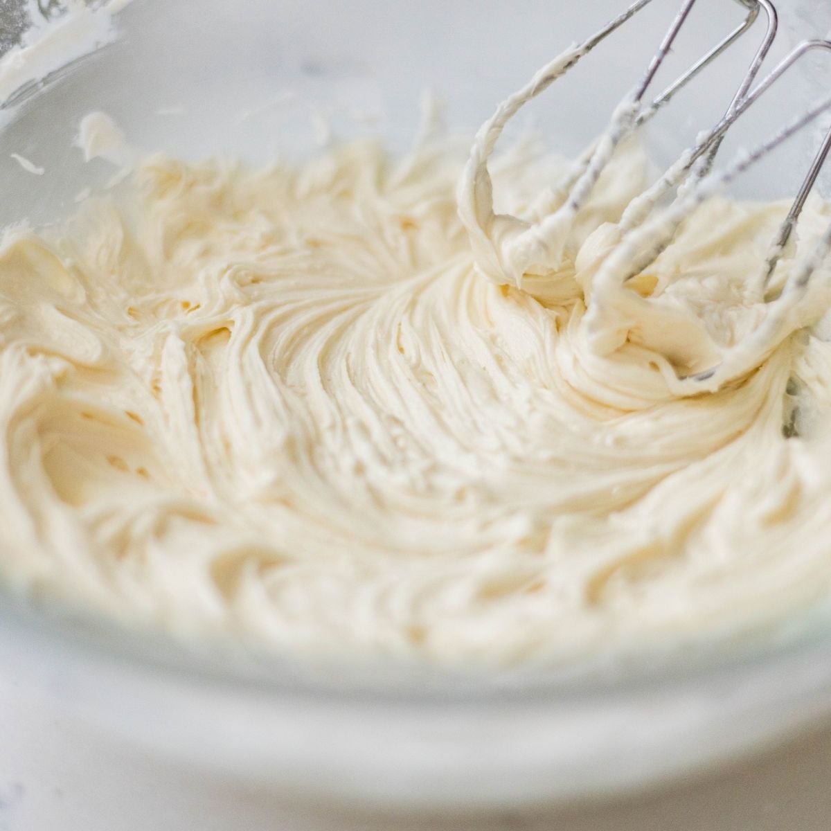 Fluffy vanilla buttercream frosting being whipped in a glass bowl with electric mixer beaters.