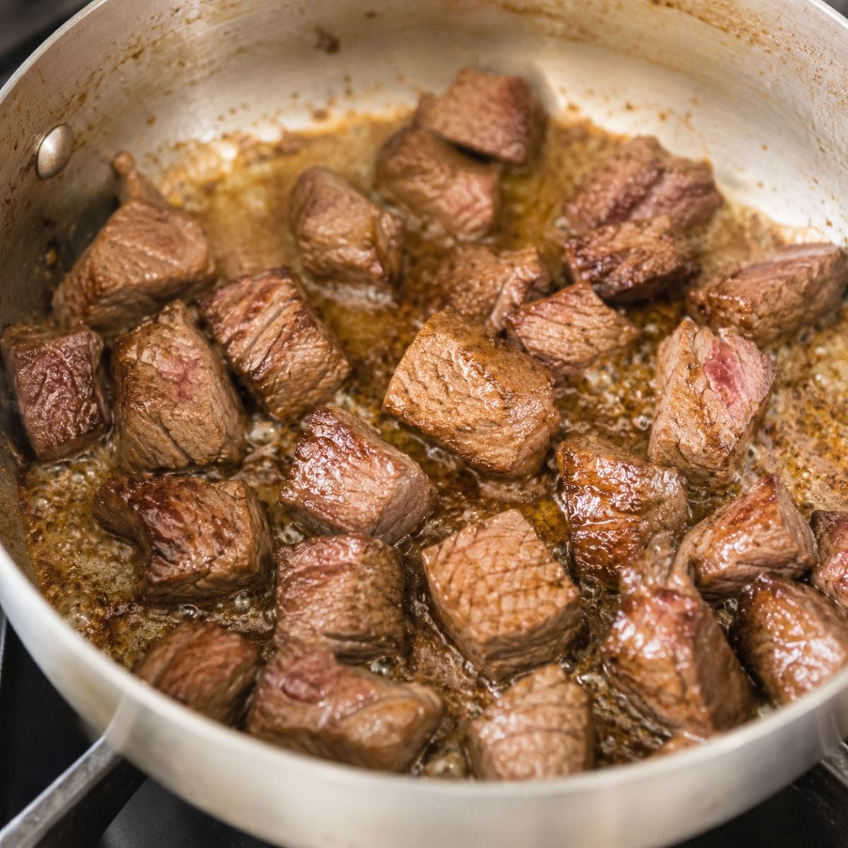 Beef cubes searing in a stainless steel pan until browned and caramelized on the outside.
